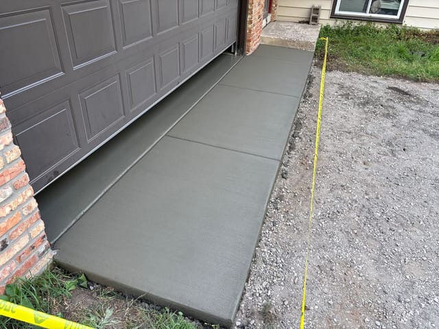 Freshly poured concrete apron and sidewalk section along a residential garage, cordoned off with yellow caution tape while curing.