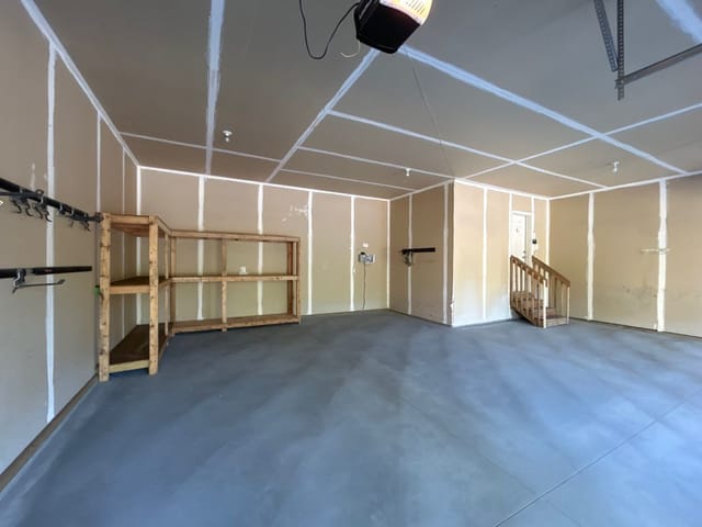 Clean residential garage interior with a newly finished gray concrete floor. Wooden storage shelves, wall-mounted hooks, and a staircase to the house line the walls, while the smooth concrete surface reflects light from the open garage door.