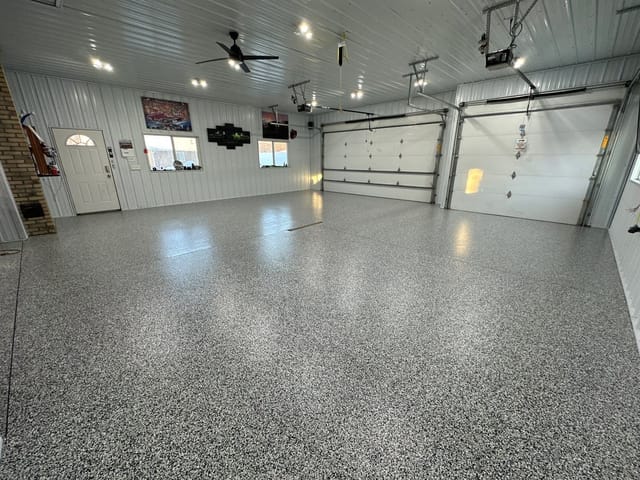 Spacious garage interior with glossy speckled epoxy flooring, white paneled walls, two large overhead doors, ceiling lights, a fan, and wall-mounted car decor.
