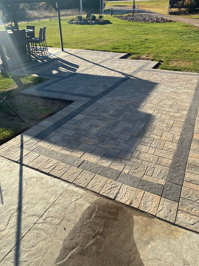 Sunlit backyard patio with textured pavers, dark accent borders, green lawn, and a covered outdoor dining area casting shadows.