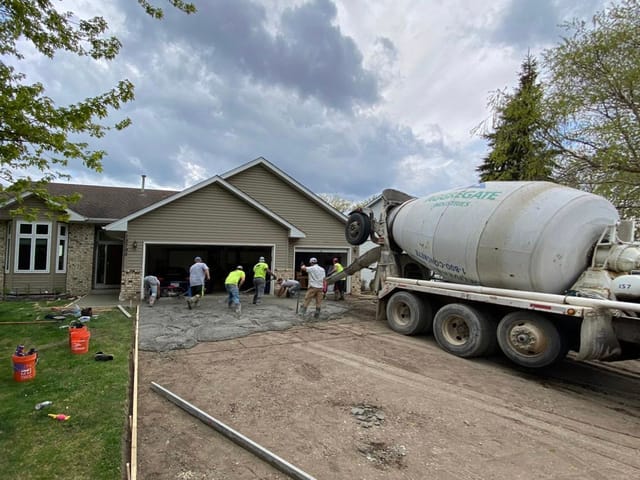 Crew pouring a concrete driveway replacement in front of a garage, with a cement truck on site.