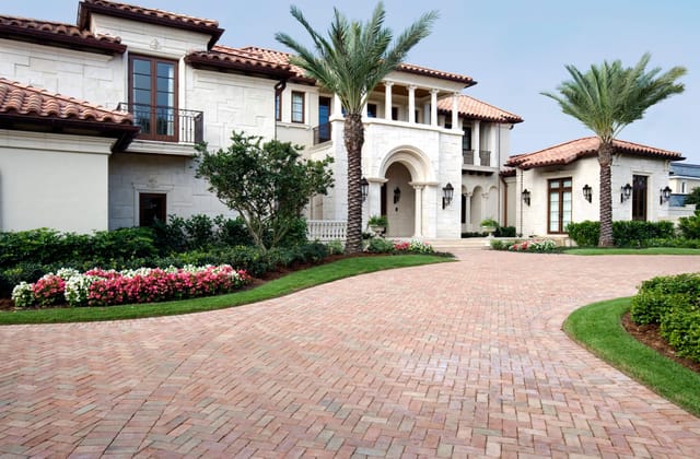 Elegant Mediterranean-style luxury home with a grand arched entryway, tall columns, red tile roof, and a wide brick driveway bordered by manicured landscaping, palm trees, and colorful flower beds.